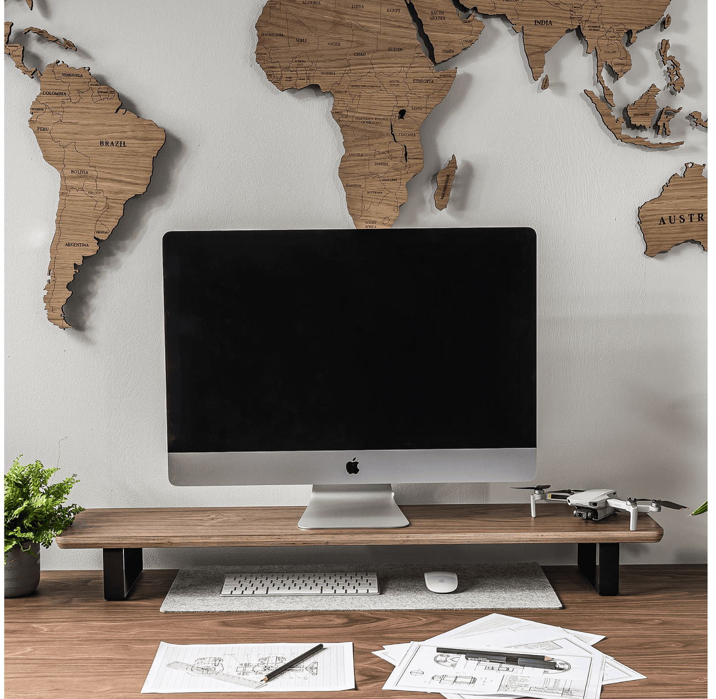 Wooden monitor stand holding an iMac, with a keyboard, mouse, potted plant, and drone on a desk, world map wall art in the background