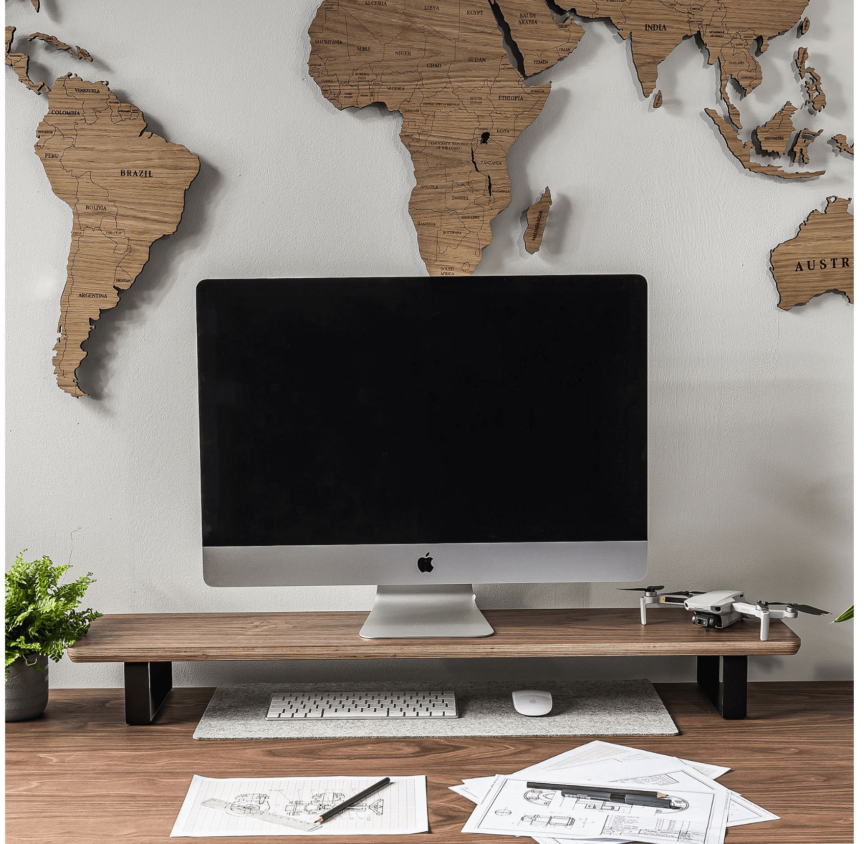 Wooden monitor stand holding an iMac, with a keyboard, mouse, potted plant, and drone on a desk, world map wall art in the background