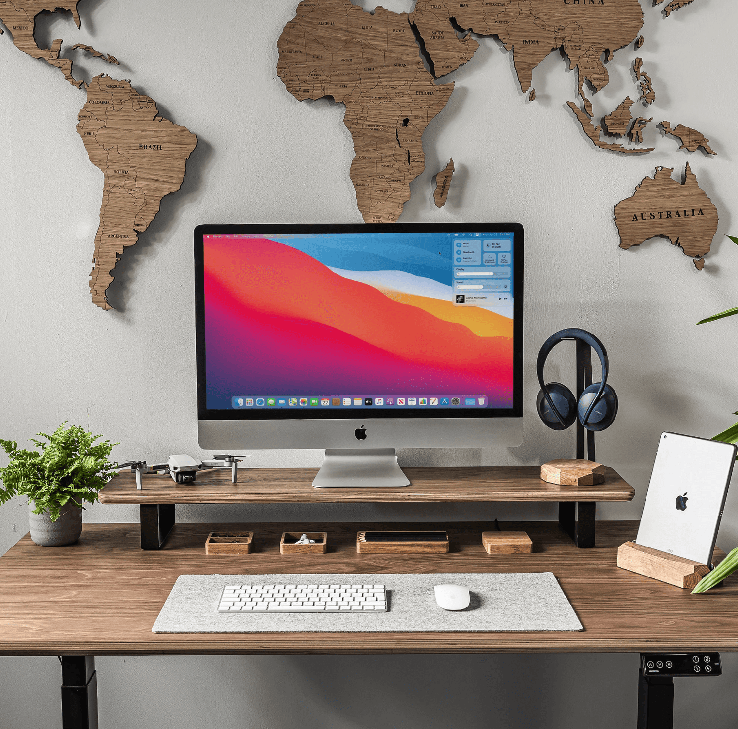 Wooden monitor stand with storage, holding an iMac, drone, potted plant, headphones, and iPad on a wooden desk, world map wall art in the background.
